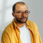 Stylish portrait of a man in glasses wearing a yellow shirt against a bright white background with natural shadows.